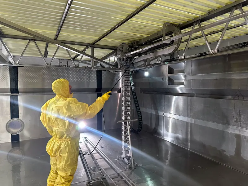 Mitarbeiter im gelben Schutzanzug reinigt mit einer Sprühlanze die Mechanik in einem leeren Edelstahlbecken