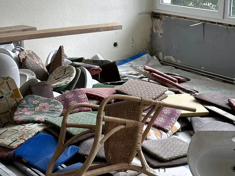 Raum bei einer Haushaltsauflösung mit gestapelten Stuhlkissen, einem Korbstuhl und einem Waschbecken