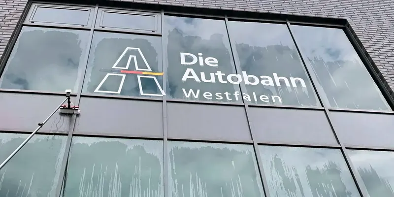 Teleskopstange mit Reinigungsbürste an einer großen Glasfassade mit dem Logo „Die Autobahn Westfalen“, auf den Scheiben laufen Wasserspuren ab
