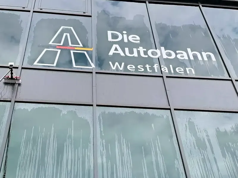 Glasfassade mit dem Logo „Die Autobahn Westfalen“ während der Reinigung, auf den Scheiben laufen Wasserspuren ab